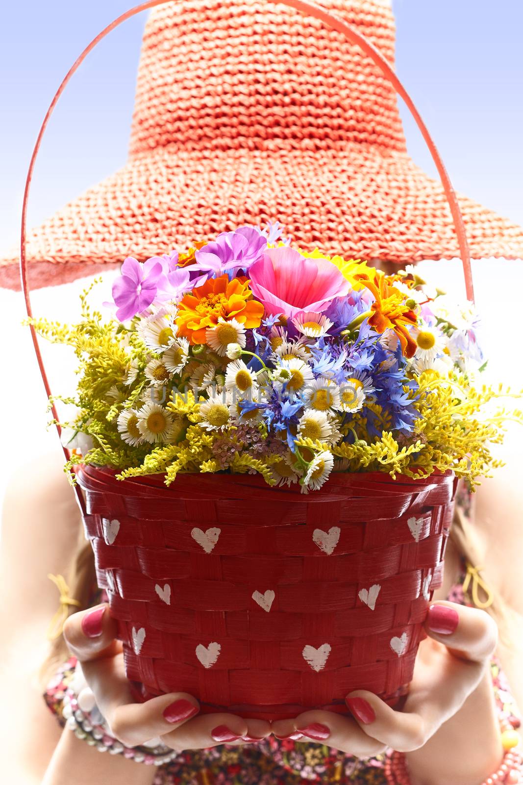 Woman with basket of wildflowers in hands. Young blonde boho girl in orange hat on summer meadow, people. Attractive joyfull, romantic style, floral sundress. Sunny day