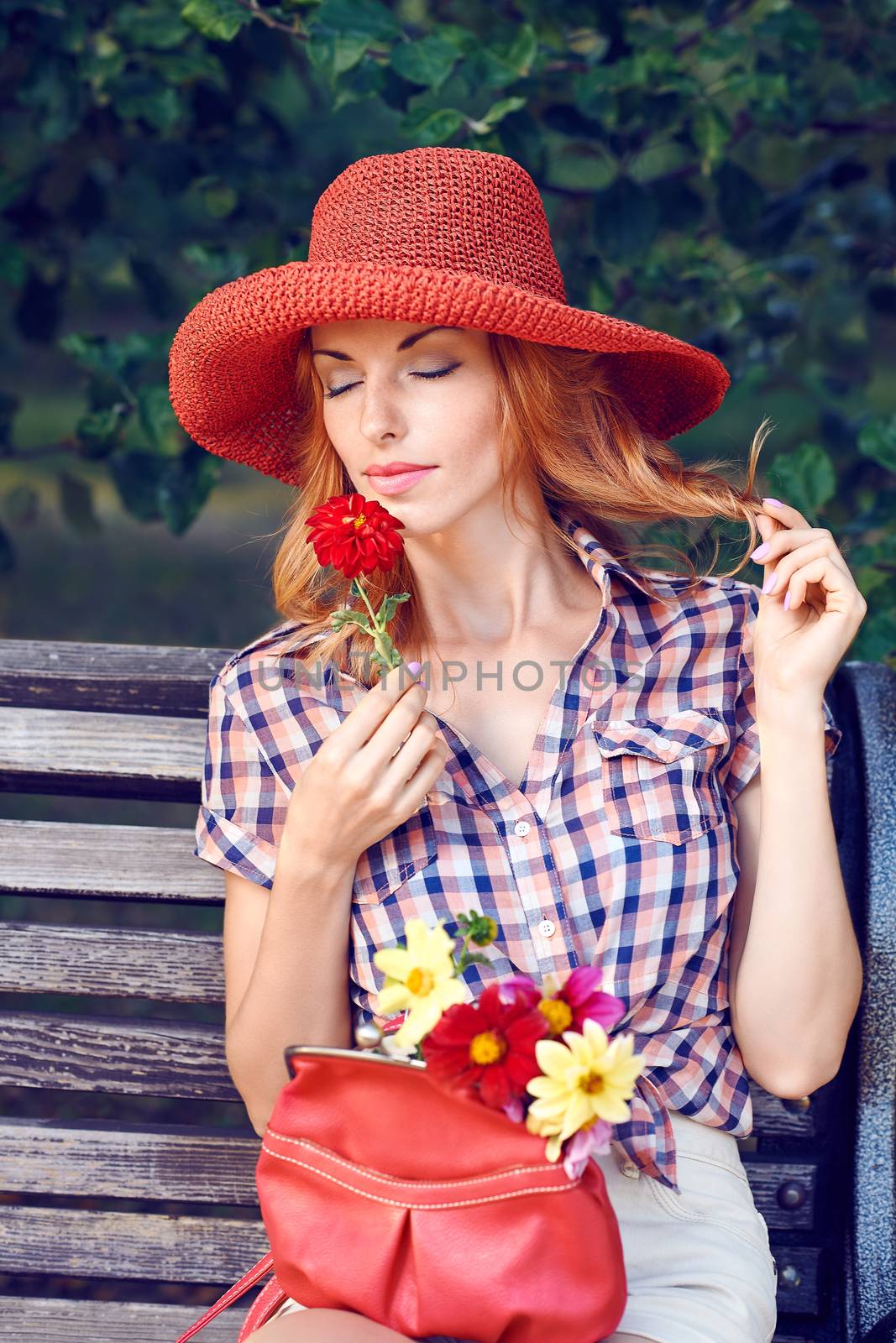Beauty portrait of stylish playful woman with closed eyes on bench in park, people, outdoors. Attractive happy girl in fashionable clothes, hat enjoying flower and nature in summer garden, lifestyle