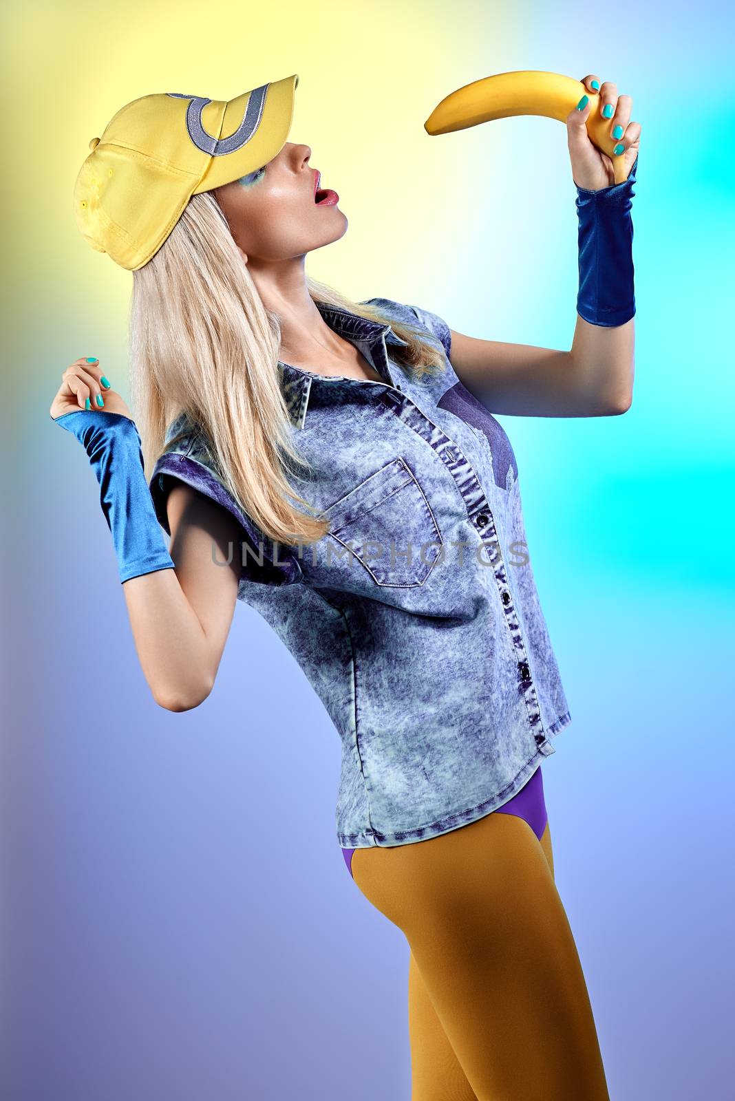 Fashion beauty sexy hipster woman in stylish clothes. Unusual creative people. Vivid girl with banana like microphone. Provocative playful blonde girl in denim, leggings, gloves, yellow cap, make up. 