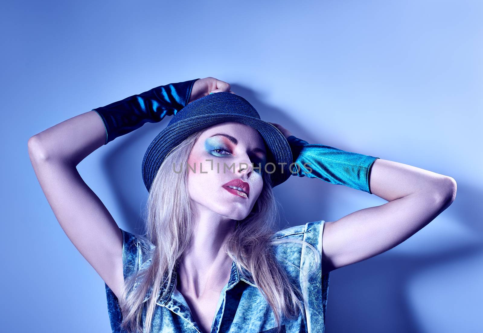 Fashion portrait of sexy beauty hipster woman in stylish clothes. Unusual creative people. Vivid girl. Provocative emotional playful blonde girl in denim shirt, gloves, hat, make up on blue, copyspace