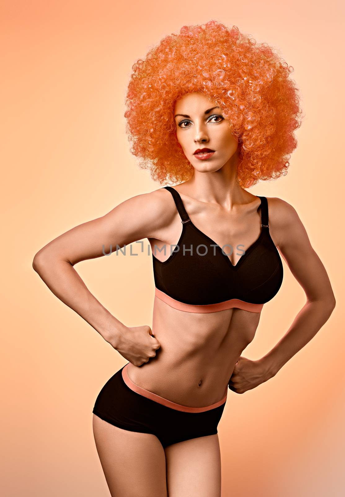 Beauty fashion. Fitness woman, unusual look. Sexy athletic body, people. Provocative attractive girl posing in trendy sport bra, shorts with afro hairstyle, orange background, copyspace, toned