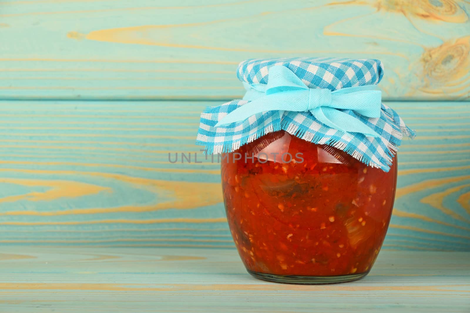 One glass jar of homemade pickled pepper, paprika and eggplant salad with checkered textile top decoration at blue painted vintage wooden surface