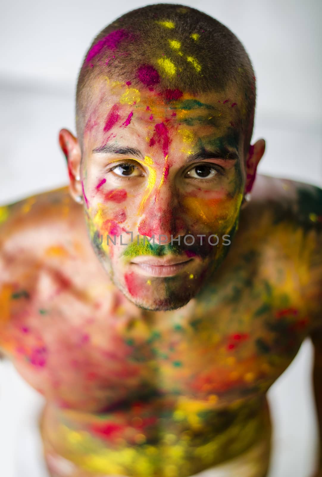 Head and shoulders shot of attractive young man shirtless, skin painted all over with bright Holi colors, looking at camera, isolated on white background