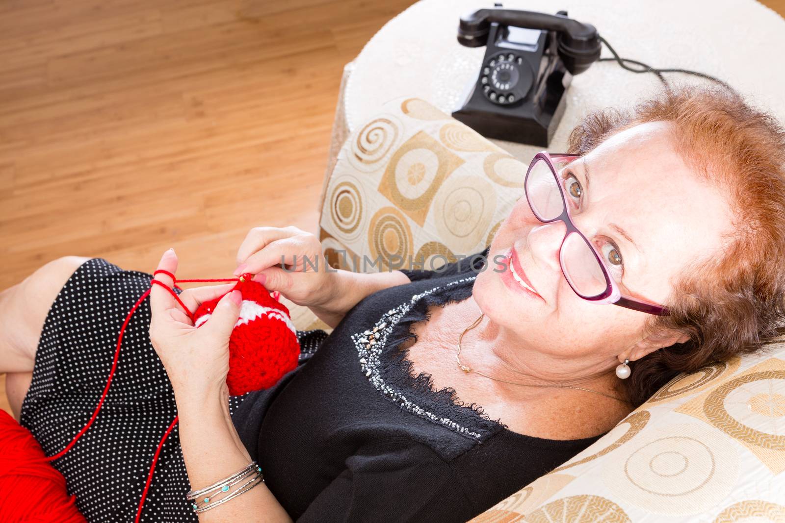 Elderly grandmother wearing eyeglasses sitting knitting in a chair looking up at the camera with a smile, old fashioned rotary telephone alongside her, high angle view
