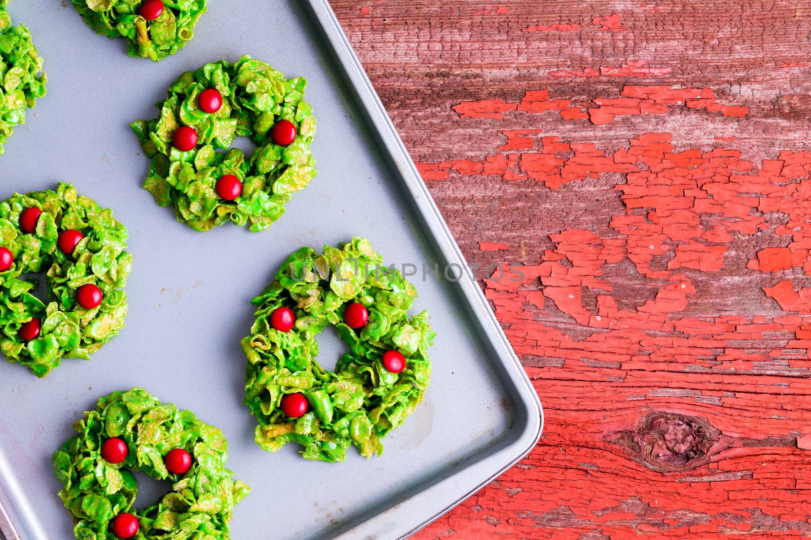 Creative colorful Xmas wreath cookies made with green cornflakes and candy pearls cooling on an oven tray on a rustic red table to celebrate the Christmas season, overhead view and copy space