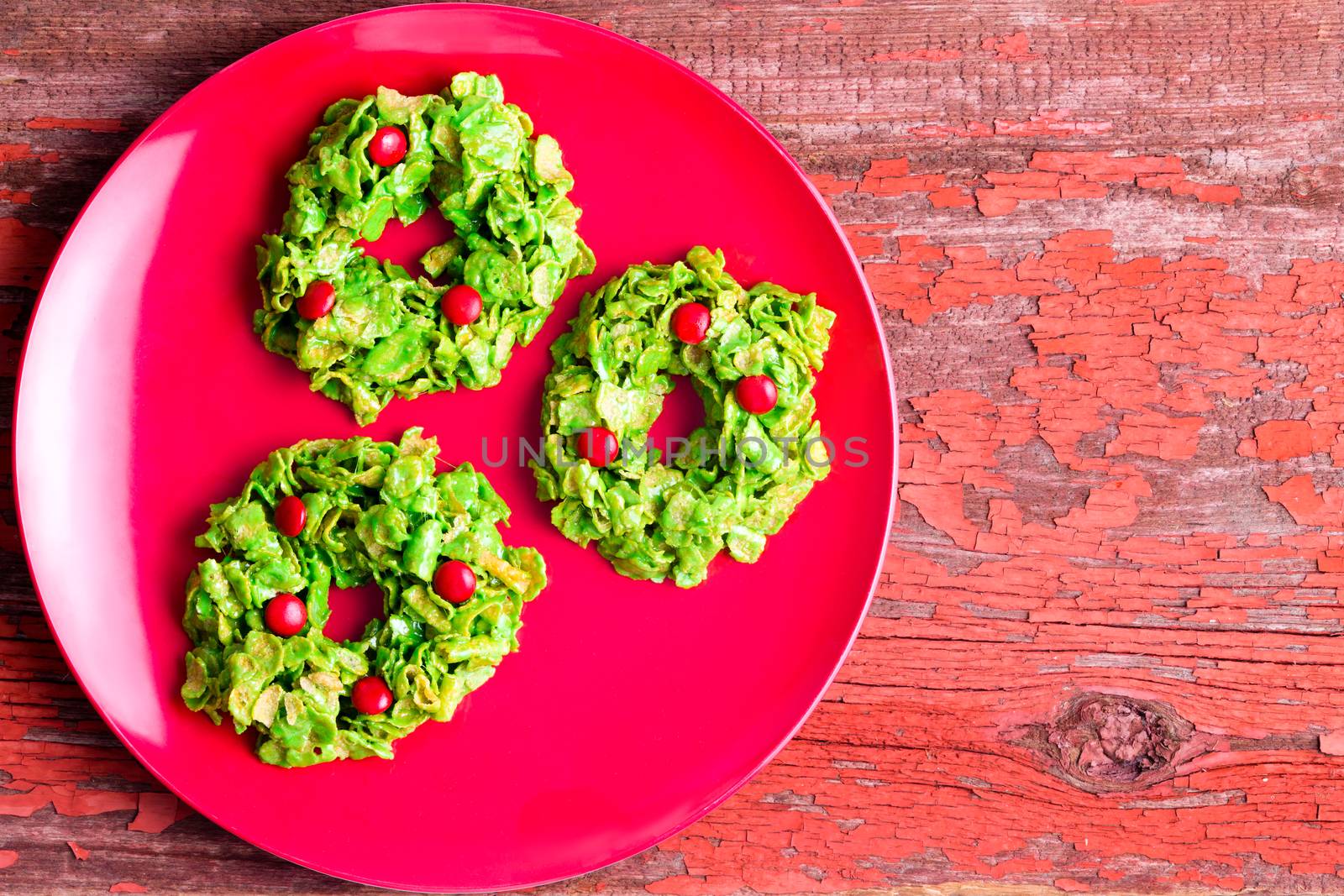 Festive colorful green Christmas cornflake wreath cookies displayed on a red plate on a cozy red rustic wooden table viewed from above with copy space