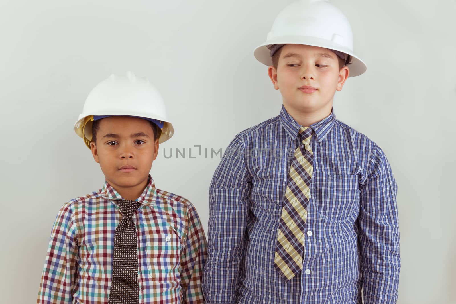 Two multiracial young aspiring tween engineers dressed in ties and hardhats standing side by side against a wall as the two friends anticipate their future careers