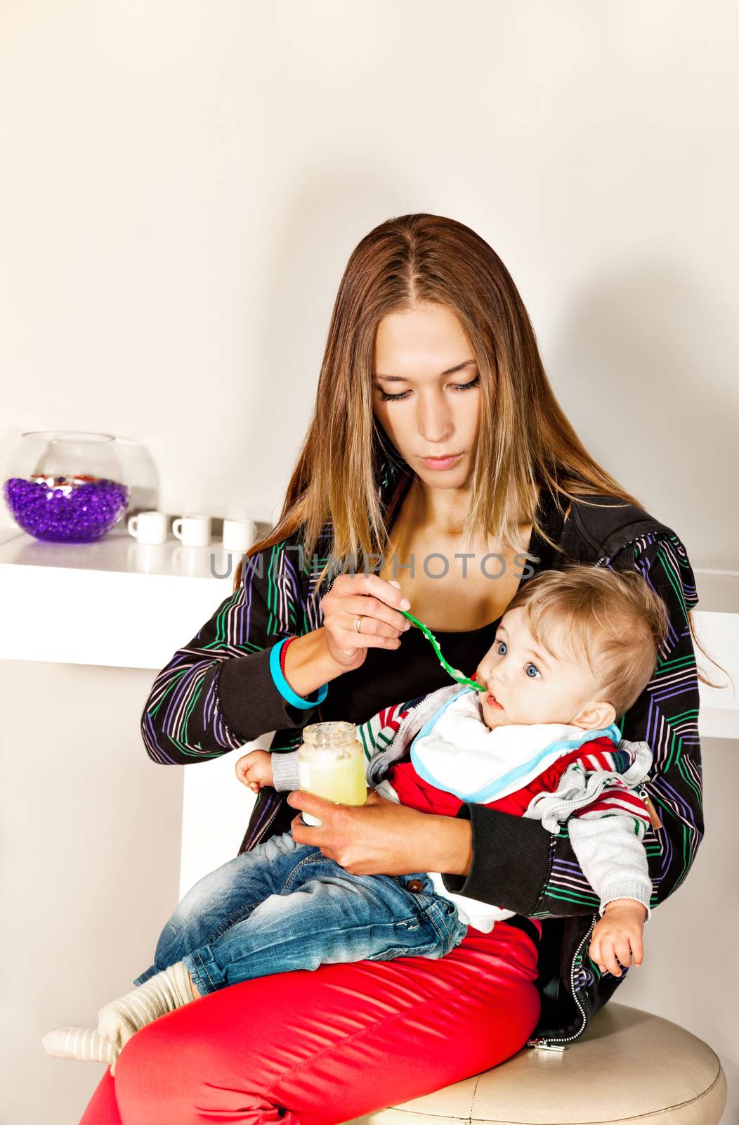 Young beautiful mother is feeding her cute little boy with a puree in modern surrounding.