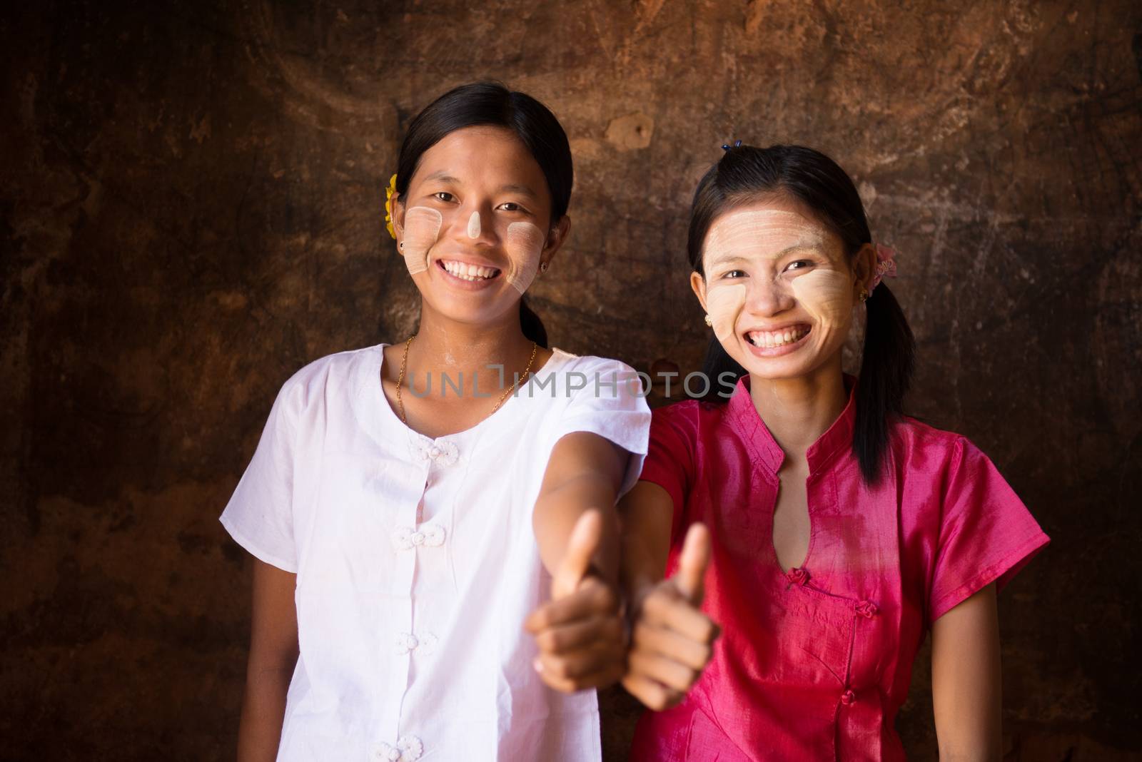 Portrait of two beautiful Myanmar girls smiling and giving thumb up, low light inside a temple.