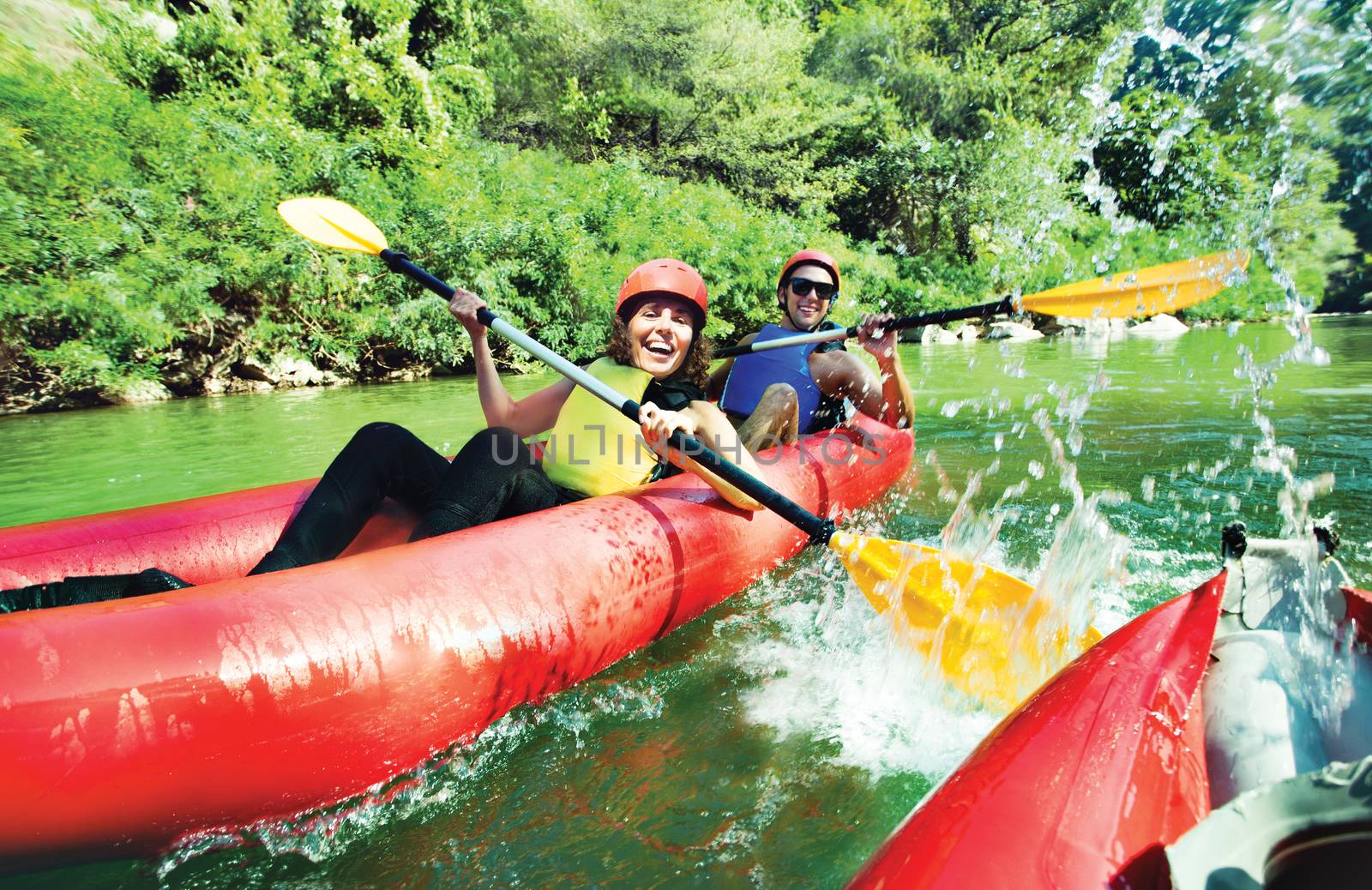 A female and a male in red inflatable canoe having fun splashing over oter with the vesals in calm waters of a river.