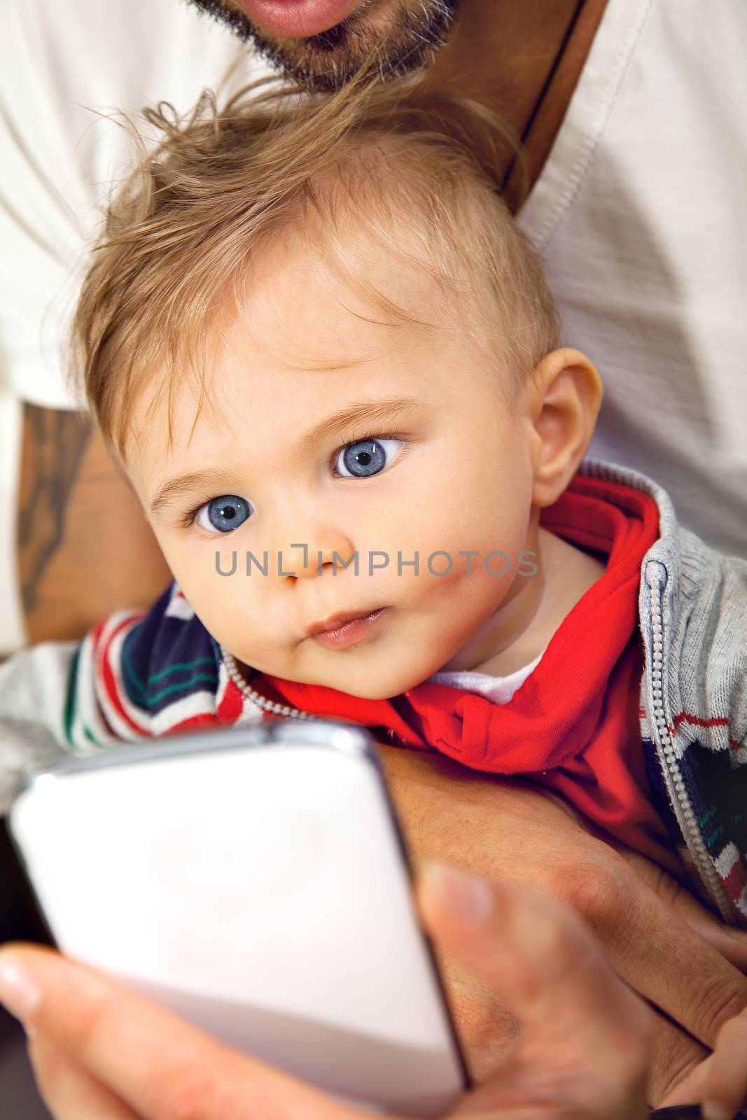 Young cute little boy is watching onto a sellphone with affection embraced by his father.