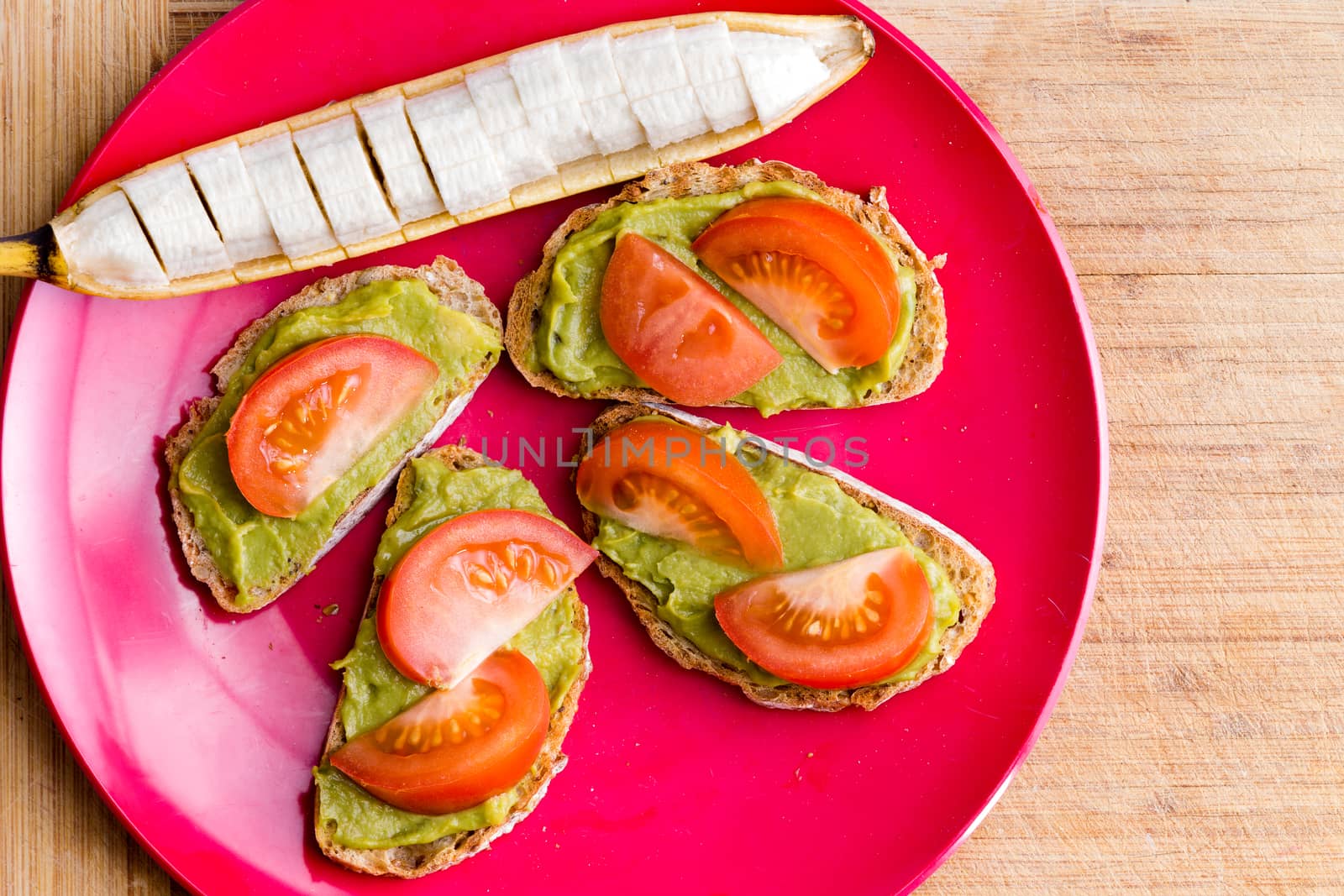 Healthy plate with sliced banana next to avocado and tomato sandwich served in red plate on wooden table
