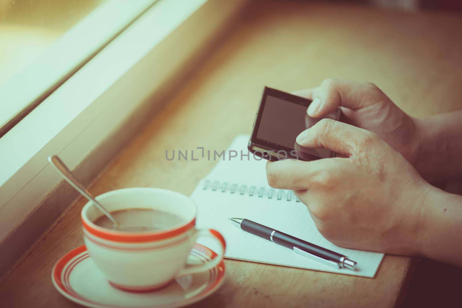 Male hand using smart phone on wood bar beside window with notebook and coffee cup in cafe in morning time with vintage filter effect