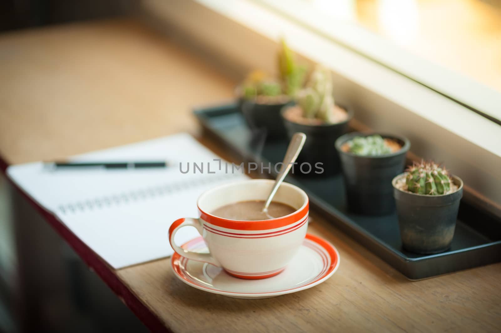 Coffee cup and opened notebook with pen on wood bar beside window at coffee shop in morning time