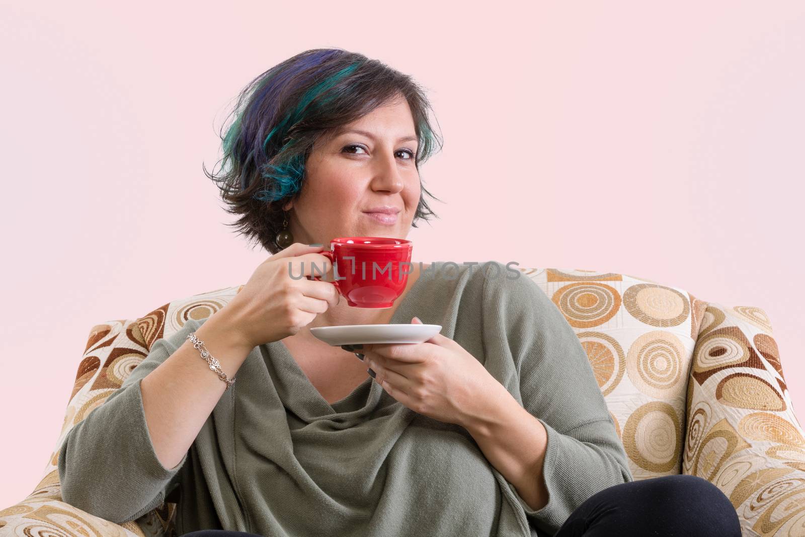 Single intelligent grinning adult female holding red coffee mug while sitting in a comfortable sofa chair