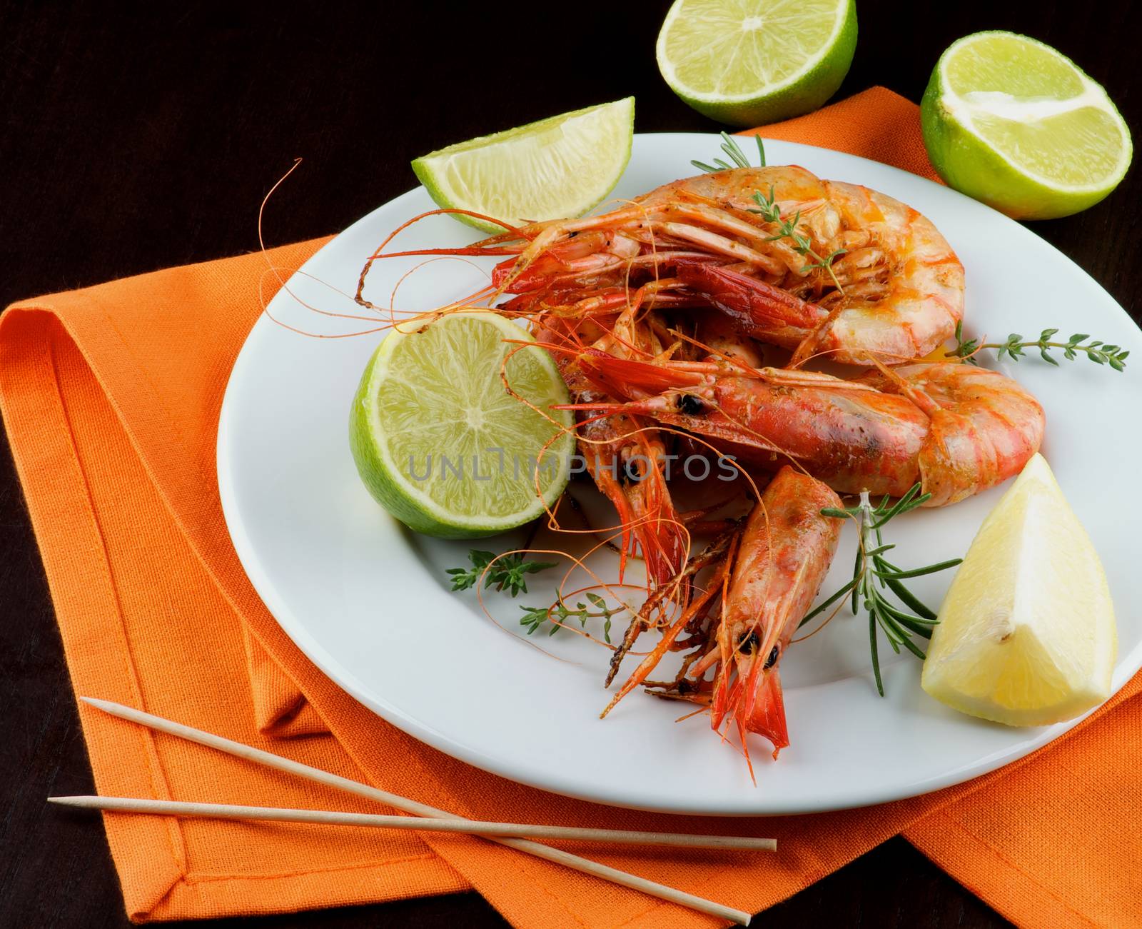 Delicious Roasted Shrimps  with Thyme, Lime and Rosemary on White Plate and Orange Napkin closeup on Dark Wooden background