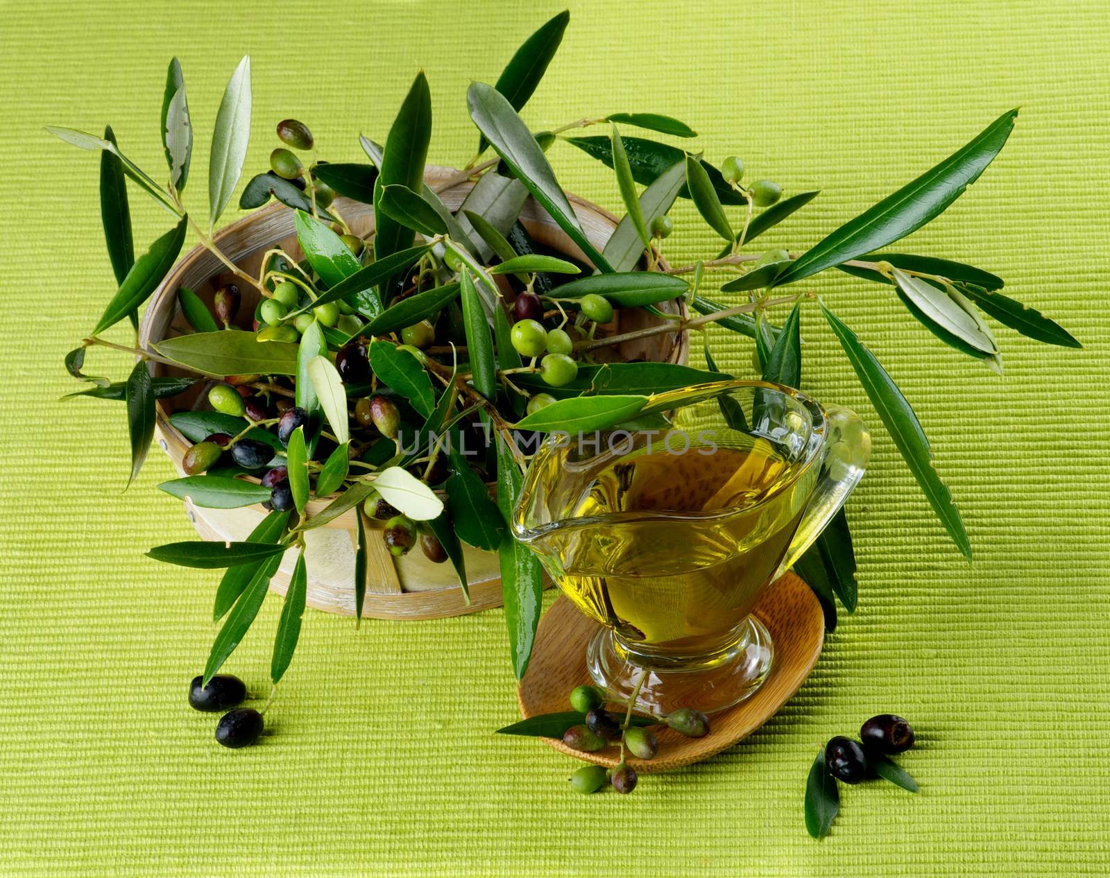 Olive Oil in Glass Gravy with Raw Green and Black Olives with Leafs in Wooden Bowl closeup on Green Textile background