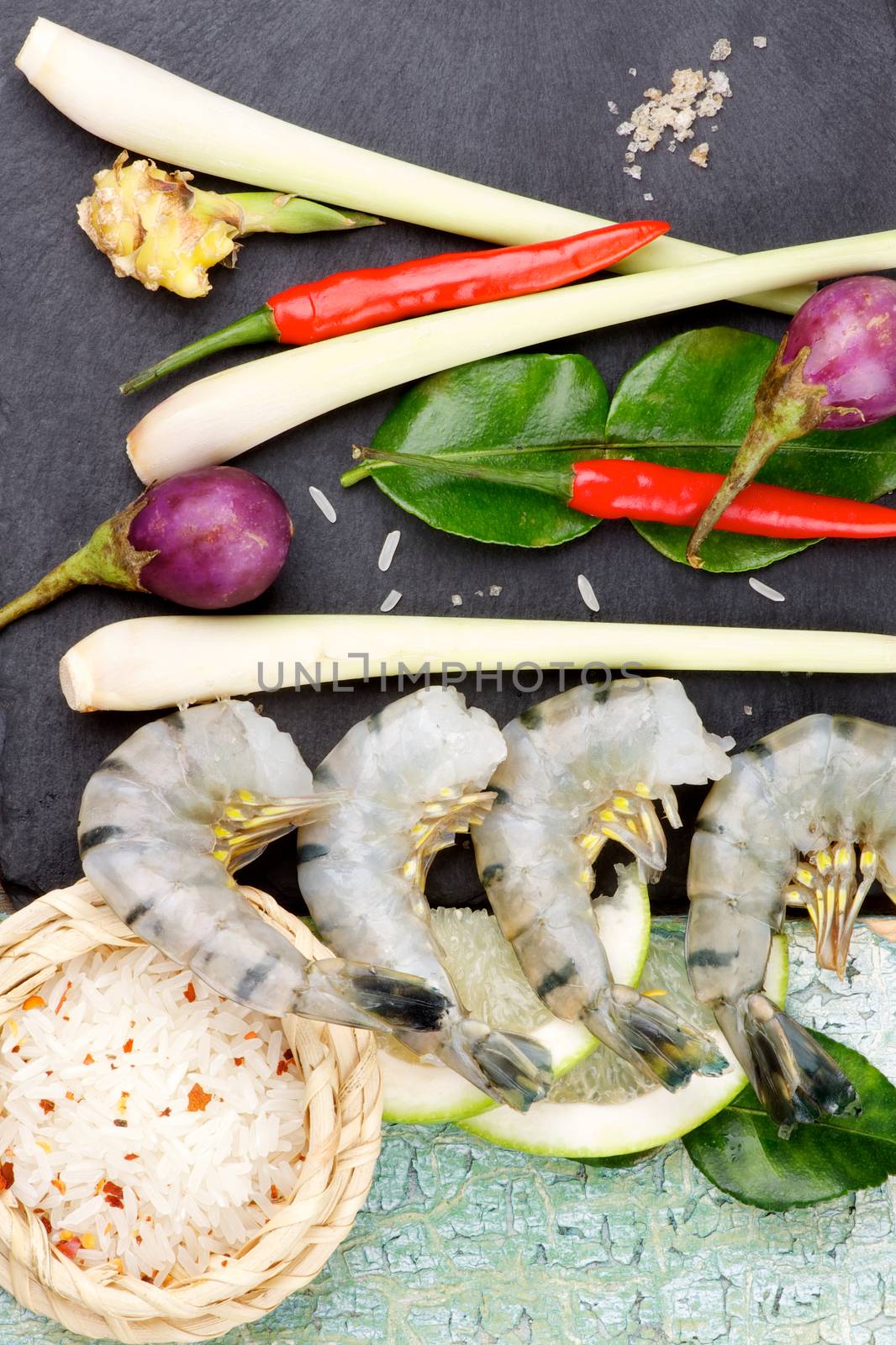 Ingredients of Thai Tom Yam Spicy Soup with Big King Shrimps, Lime, Lemon Grass, Chili Peppers and Spices, Raw Rice and Greens closeup on Slate and Wooden background. Top View