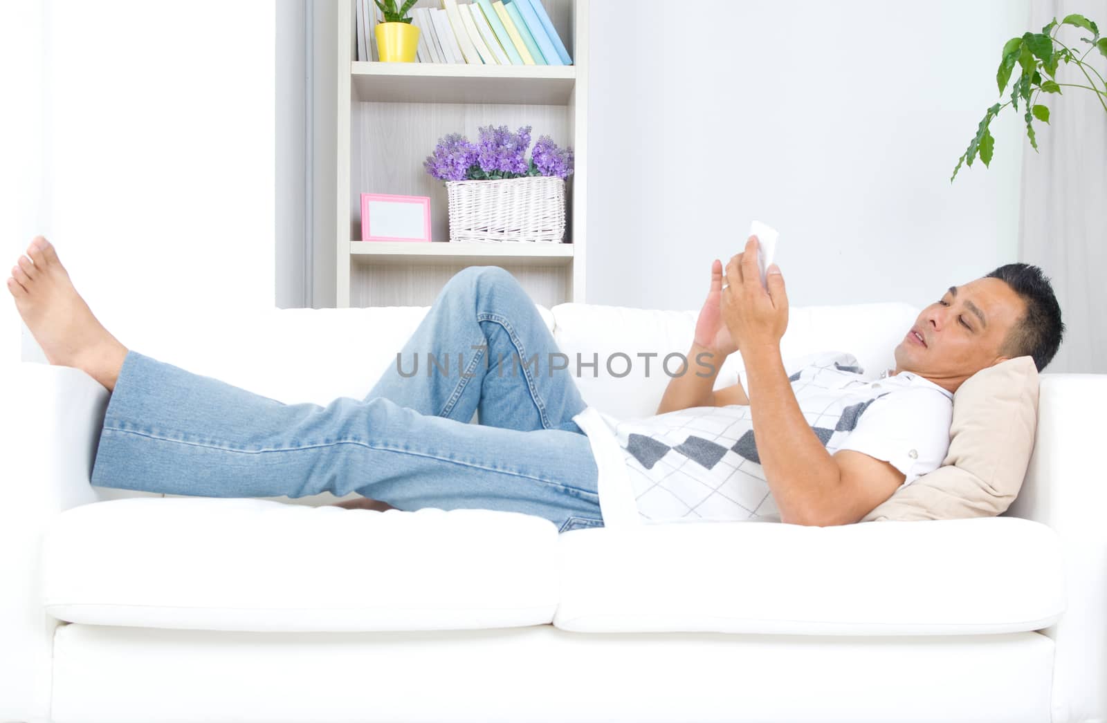 Smartphone mobile apps concept. Malay guy using smart phone. Handsome asian man relaxed and lying on sofa indoor.