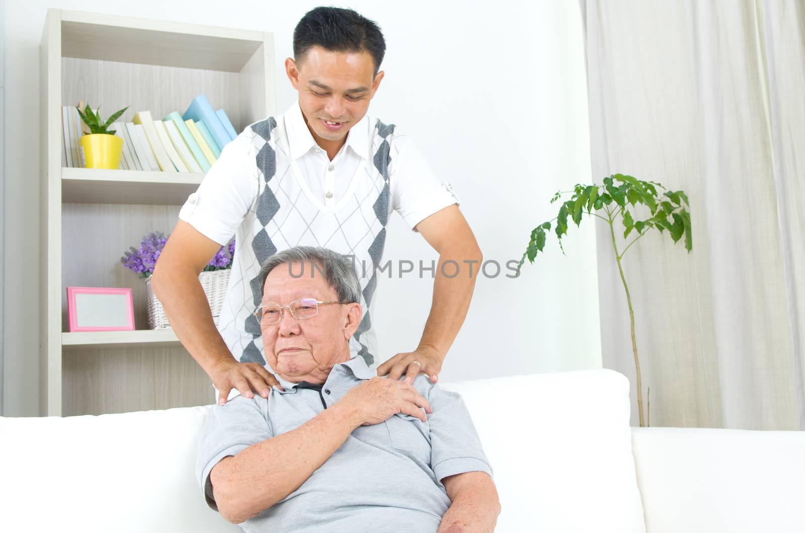 Asian old man shoulder pain, sitting on sofa , son massaging father shoulder. Chinese family, senior retiree indoors living lifestyle at home.