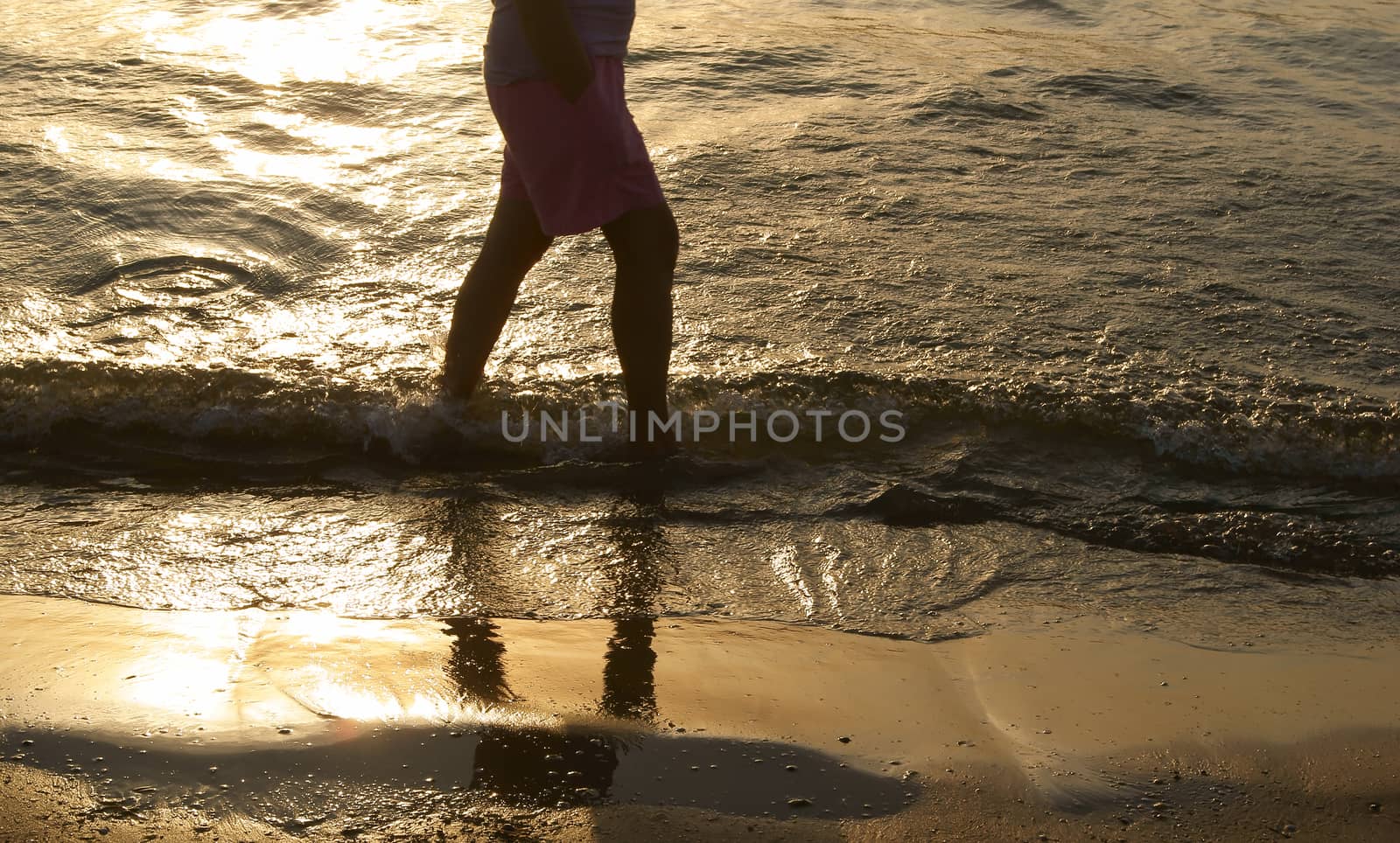 Silhouette of a male with arms in the pockets walking along the sea water at sunset