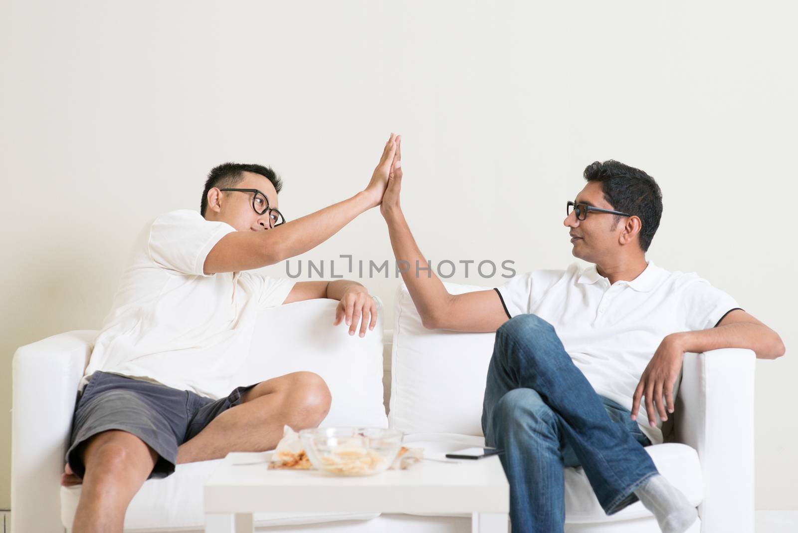 Men talk concept. Guy sitting on sofa and giving high five to friend at home. Multiracial people friendship.