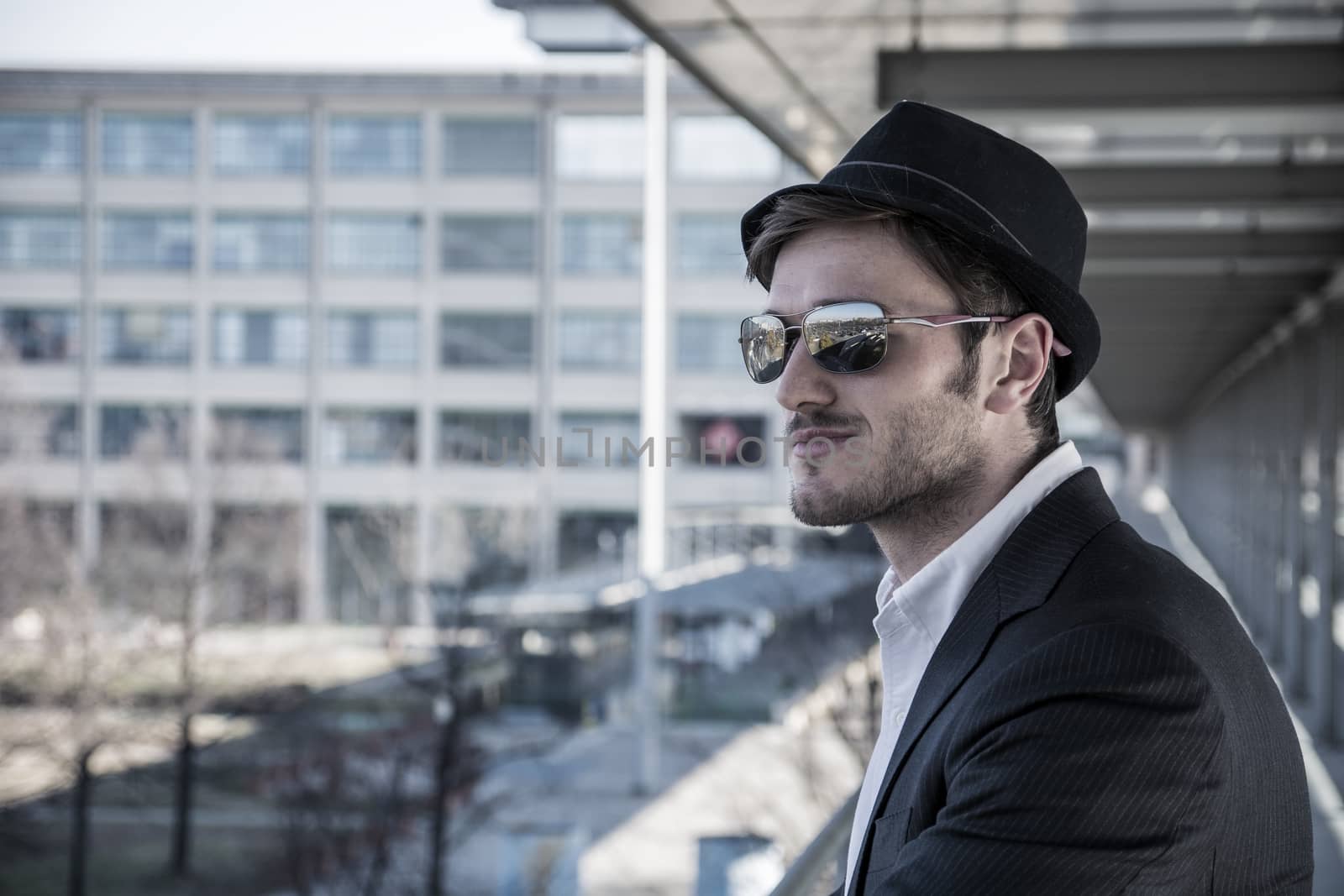 Head and Shoulders Portrait of Stylist Young Man Wearing Suit and Hat Looking to the Side Out Window While Standing on Moving Sidewalk in Building