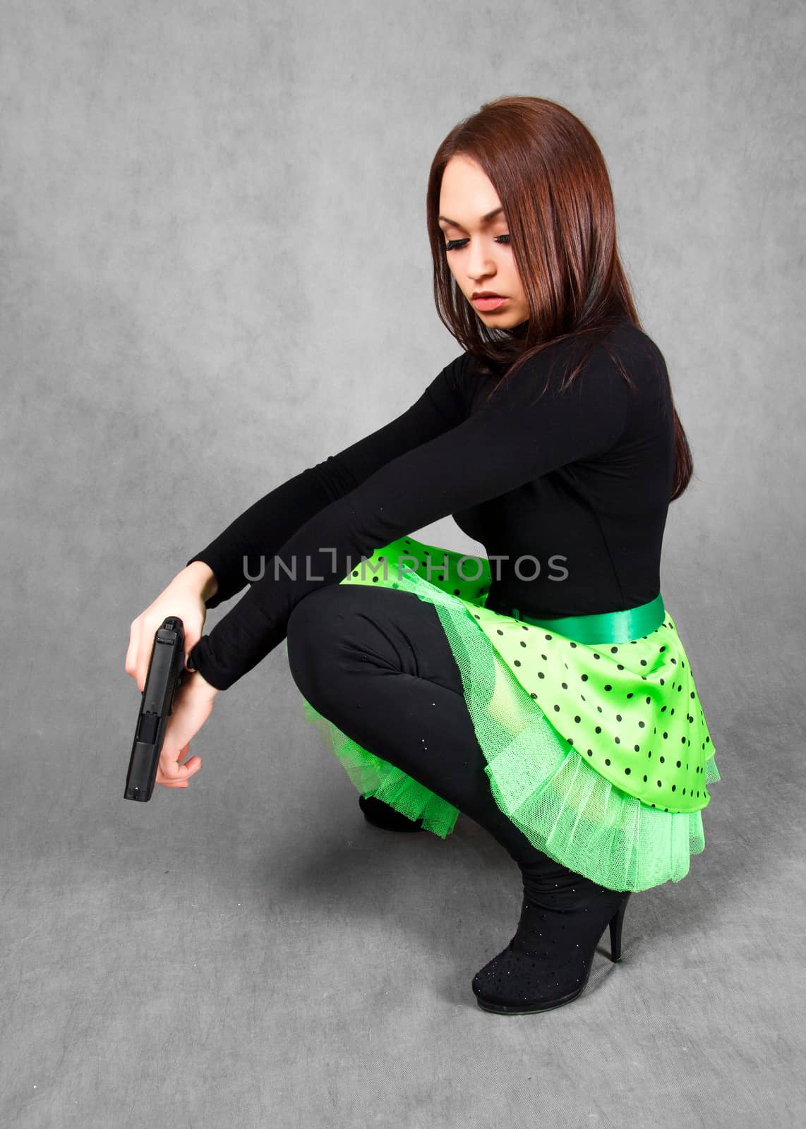 Portrait of a young attractive woman in a bright green skirt with a gun over grey background