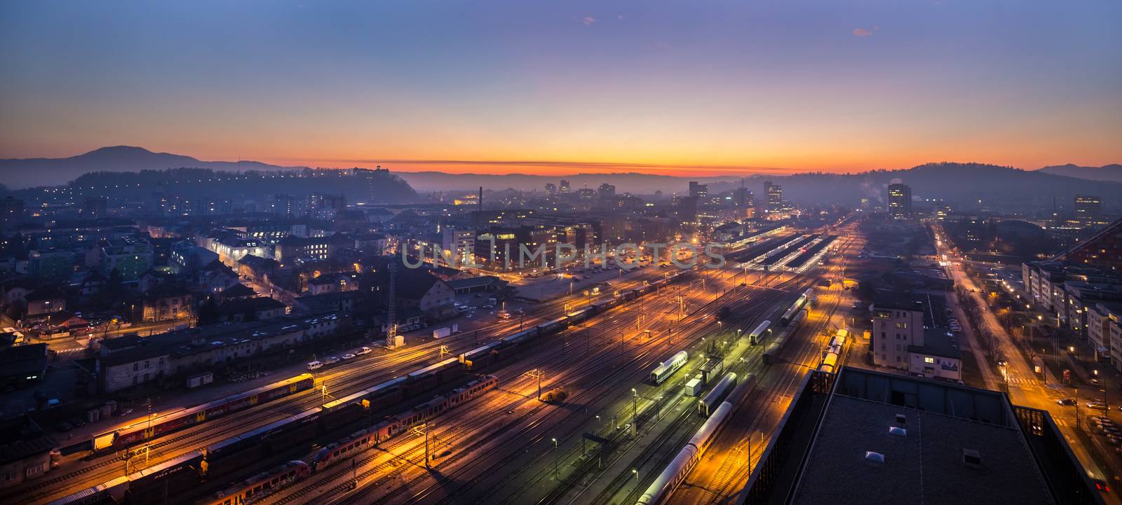 Cityscape of Slovenian capital Ljubljana at dusk. Aerial view of illuminated train tracks of a main railway station can be seen in forground.