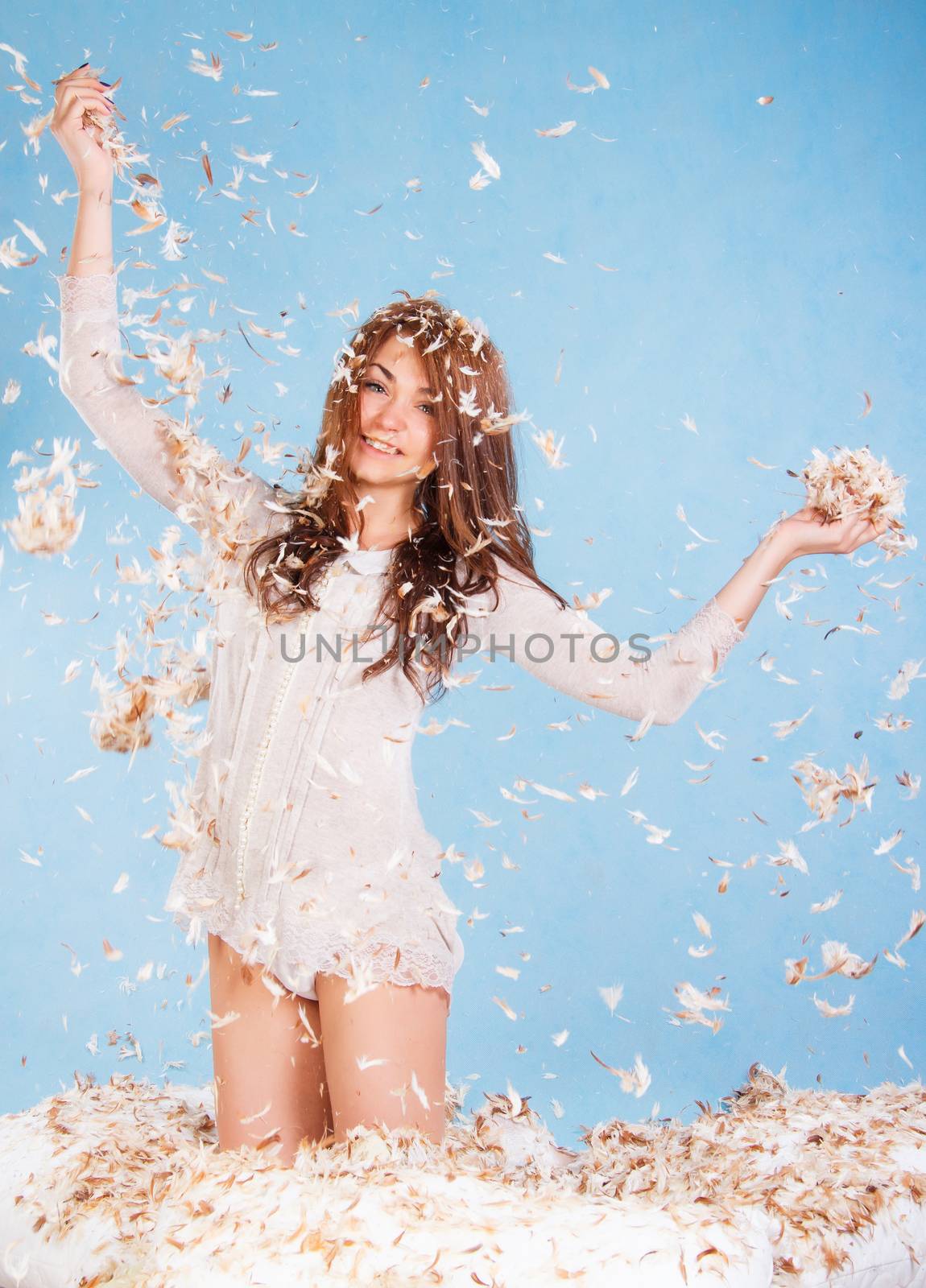 Beautiful young woman in a bed dropping the feathers over blue background