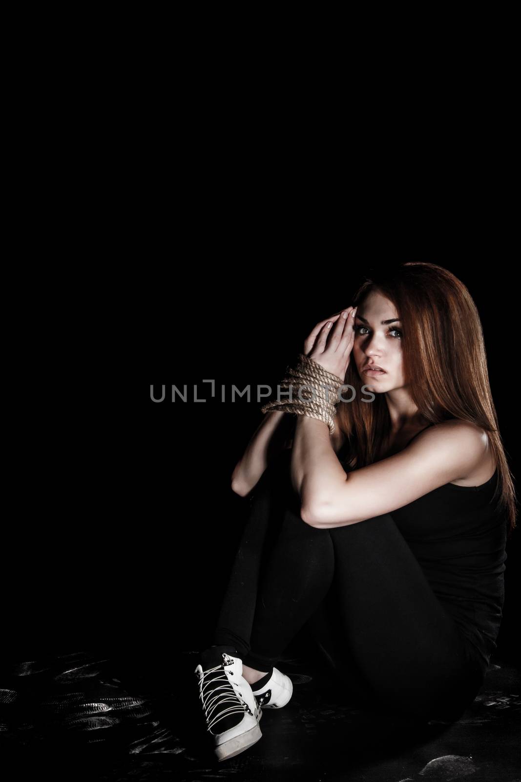Beautiful young woman with tied arms sitting on the floor over black background