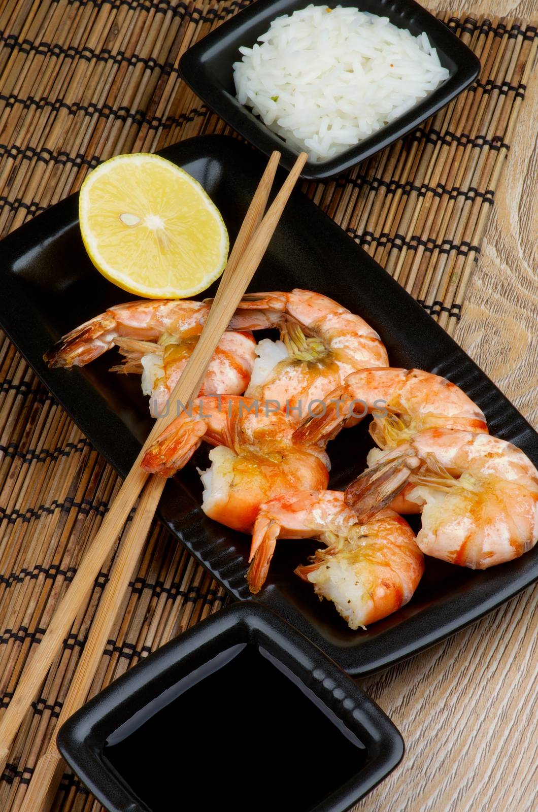 Delicious Roasted Shrimps with Lemon and Chopsticks, Soy Sauce and Boiled Rice in Square Bowls on Straw Mat background in Asian Style