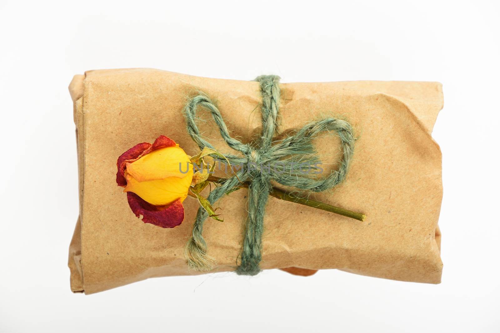 Kraft paper wrapped small romantic holiday gift with green burlap jute rope bow and dried up rose isolated on white background