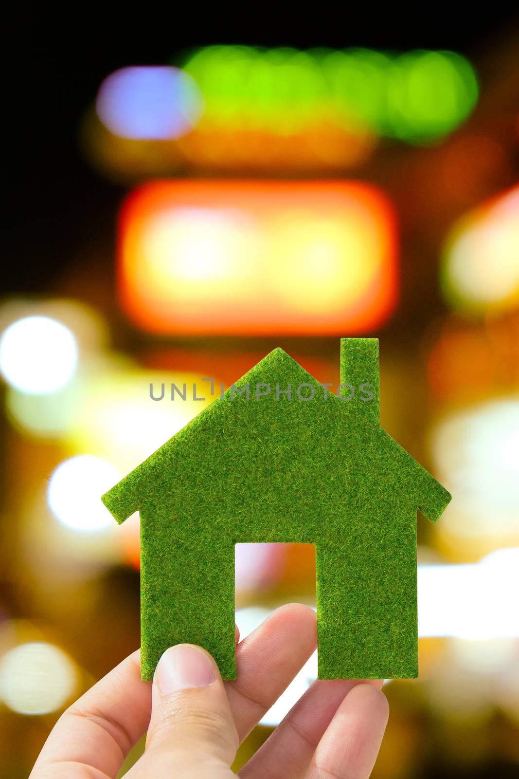 Hand holding small green eco house icon with defocused city night light background, green energy concept