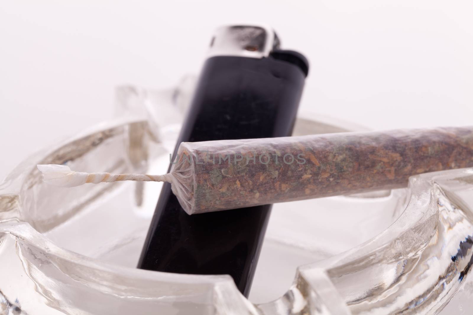 Close up of marijuana joint made with translucent rolling papers, plastic baggy of dried marijuana, black lighter and pipe on white background