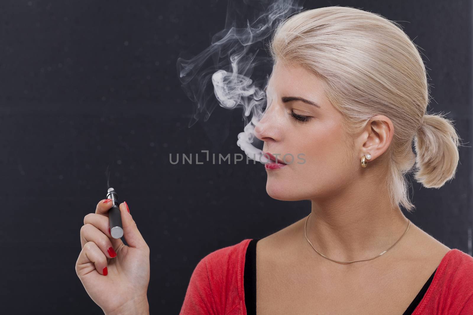 Stylish blond woman smoking an e-cigarette exhaling a cloud of smoke with her eyes closed in enjoyment, profile view on a dark background