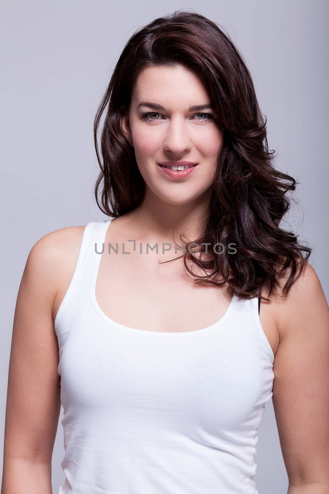 Smiling attractive woman with a lovely warm friendly smile and shoulder length curly brown hair looking at the camera over a grey background