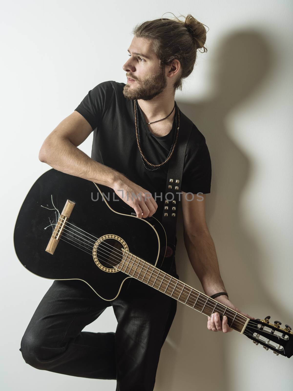 Photo of a young attractive man with long hair and beard leaning against the wall holding an acoustic guitar.

