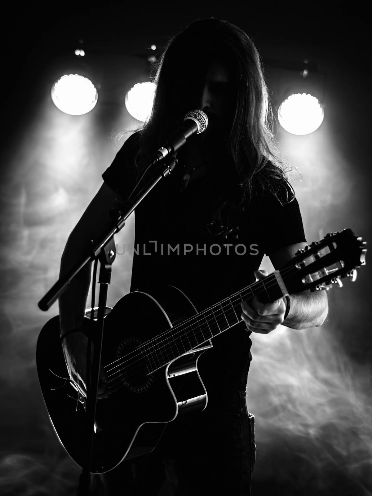 Photo of a backlit young man with long hair in silhouette playing an acoustic guitar on stage.

