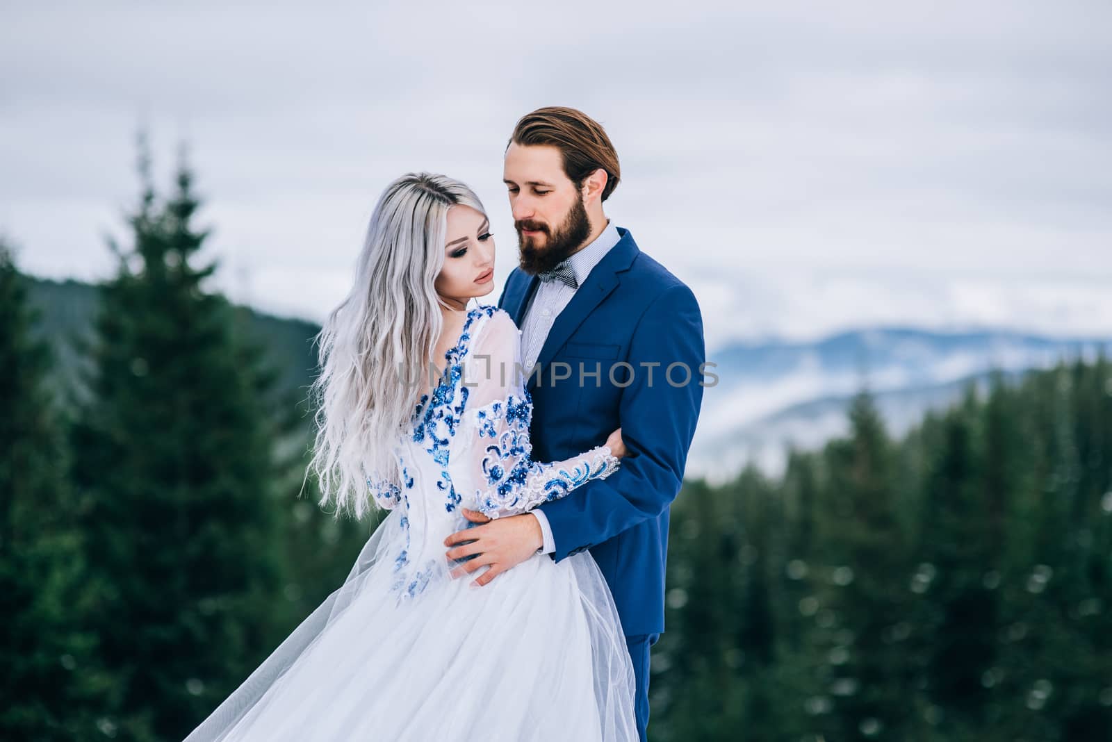 groom in a blue suit and bride in white, embroidered with blue pattern, dress on a background of green pine forests in the mountains of the Carpathians