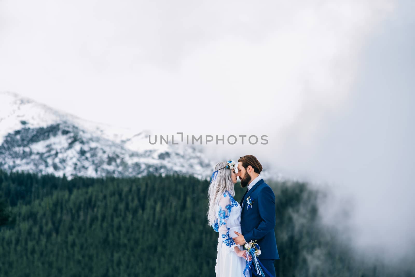 groom in a blue suit and bride in white, embroidered with blue pattern, dress on a background of green pine forests in the mountains of the Carpathians