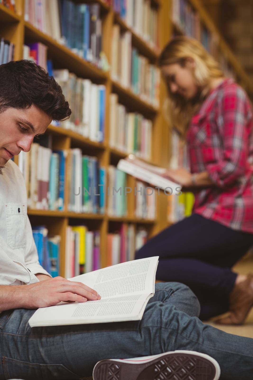 Smiling classmates reading book while leaning on bookshelves in library