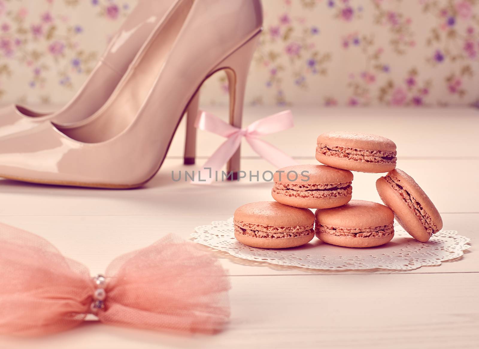 Woman essentials fashion accessories. Macarons french dessert, luxury beige shoes high heels, bow. Creative glamor wedding set, vanilla wood, floral background. Romantic, still life. Retro vintage 
