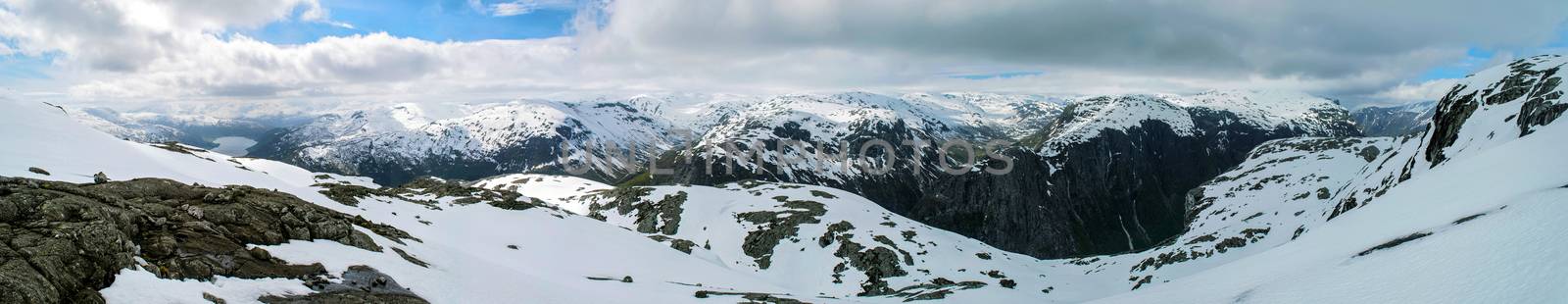 Wide panorama of snowy mountains of Norway. Cliffs stones, snow, lake. Cloudy weather, some sunlight.