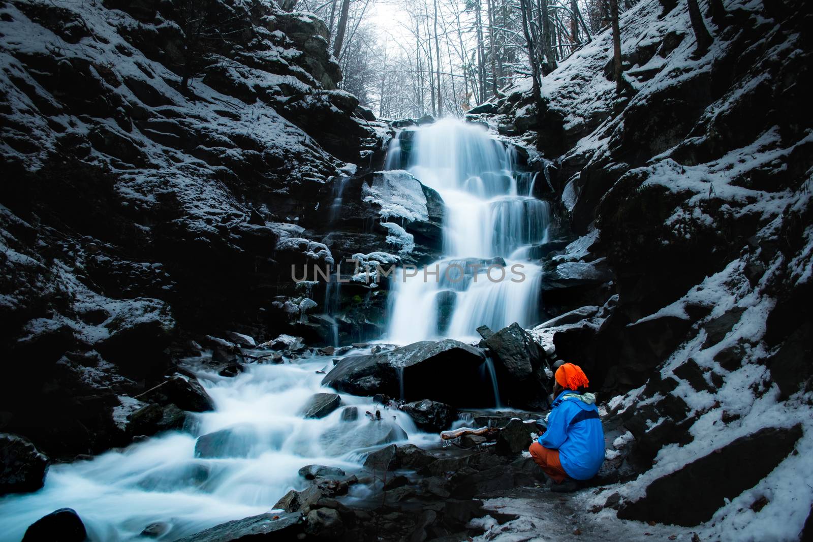 Girl in sportswear sits in front of a waterfall. Long exposure. Water is blurred. Winter. Cold conditions. Blue water.