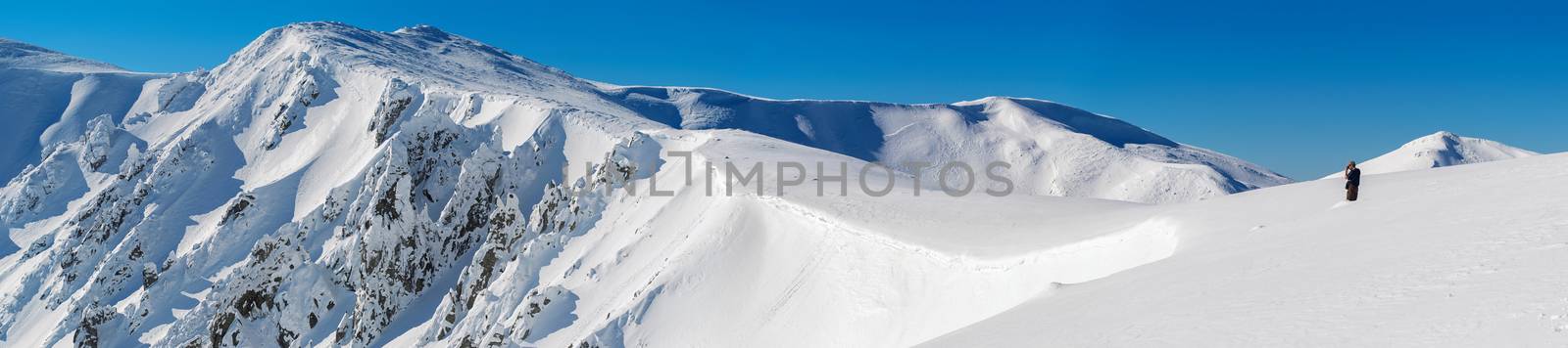 Panorama of snow-covered rocks of the mountain range. A man standing in the snow at the edge of the slope. The sky is clear, sunny. Winter. Ukraine