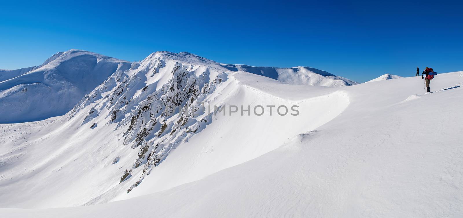 Panorama of snowy mountain peaks. Sharp rocks on a steep slope. Two tourists. The sky is clear, sunny. Winter. Ukraine