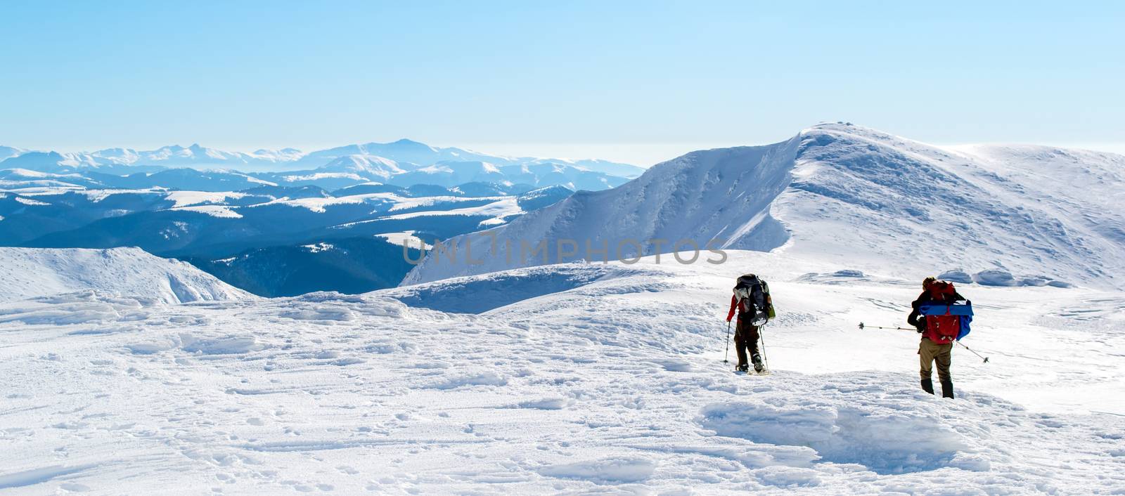 Two tourists walking on snow-covered mountain ridge towards the summit. The sky is clear, sunny. Winter. Ukraine