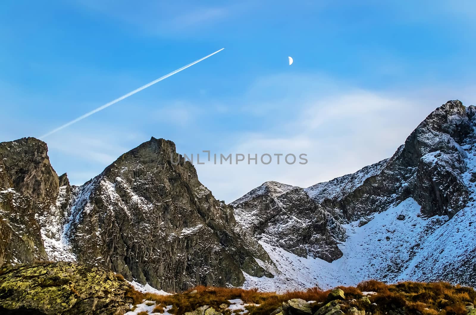 Plane flies over the rocky peaks of high mountains. Leaving a trail thru blue sky with the moon. Autumn landscape. Romania