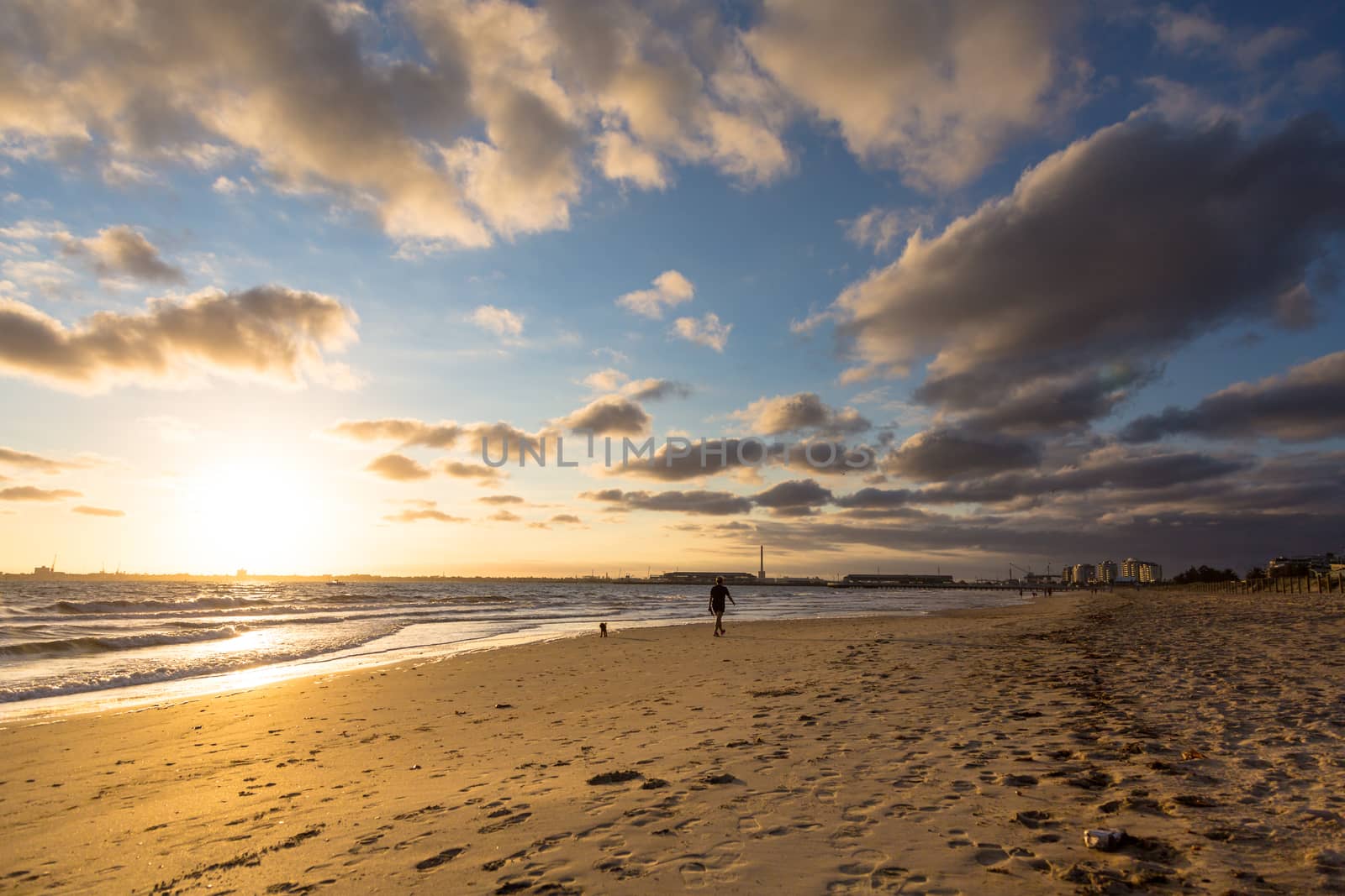 Sun setting on an urban beach on a cloudly afternoon, sand in the foreground with a person walking their dog.